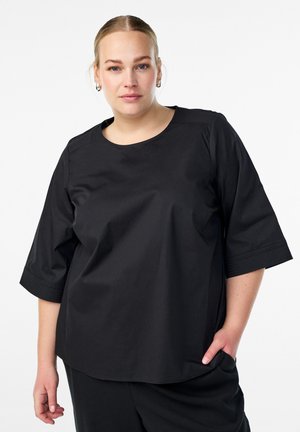 Žena s plavom kosom u crnoj, širokoj majici i crnim hlačama, stoji s jednom rukom u džepu pred bijelom pozadinom.