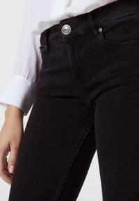 Vaqueros de mezclilla negra con una textura suave, cintura alta y cierre de botón plateado, combinados con una blusa blanca cuyo puño es visible en la muñeca.