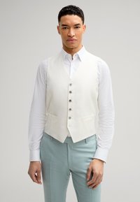 Weißer, strukturierter Weste mit fünf silbernen Knöpfen, kombiniert mit einem weißen Hemd und hellblauen Hosen. Fitted-Design, keine sichtbaren Muster oder Akzente.