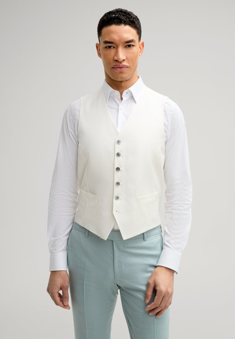 Weißer, strukturierter Weste mit fünf silbernen Knöpfen, kombiniert mit einem weißen Hemd und hellblauen Hosen. Fitted-Design, keine sichtbaren Muster oder Akzente.
