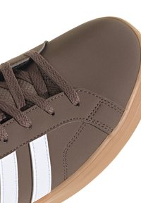 Zapatilla marrón con rayas blancas laterales, cordones marrones y suela de goma marrón clara, mostrando detalles delanteros y laterales.