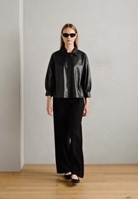 Zwarte satijnen blouse met knopen en opgeblazen mouwen, gecombineerd met zwarte wijduitlopende broek en zwarte flats, tegen een neutrale achtergrond.
