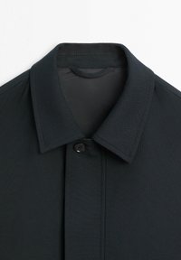 Kołnierz ciemnoszarego płaszcza z jednym czarnym guzikiem zapiętym pod klasycznym spiczastym kołnierzem na jednolitym tle.