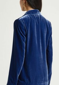 Veste en velours bleu marine avec un col classique, des manches longues et une texture lisse, dotée d'un léger éclat et d'une coupe ajustée. Vue de dos montrée.
