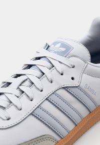 Vita lädersneakers med ljusblå detaljer, tre ränder och gummisula. Har en vadderad krage och platta snören. Texten säger "SAMBA."