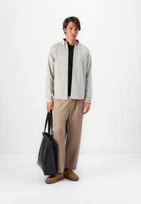 Man wearing light gray buttoned shirt over black top, beige wide-leg pants, brown shoes, holding large black tote bag, standing on white background.