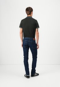 Polo shirt vert foncé avec des accents blancs, associé à un jean en denim bleu foncé. La vue arrière montre une coupe ajustée et un branding minimal sur le jean.