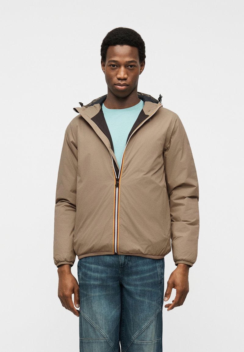 Νέος άνδρας που φοράει μπεζ ζακέτα με φερμουάρ πάνω από ανοιχτό μπλε πουκάμισο και μπλε τζιν, στέκεται μπροστά σε ένα απλό ανοιχτό φόντο.