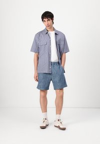 Camisa de cuadros de manga corta en azul y blanco, con dos bolsillos frontales, llevada sobre una camiseta blanca y combinada con pantalones cortos de mezclilla desgastados.