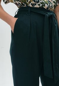 Ciemnozielone spodnie wykonane z teksturowanej tkaniny. Posiadają zwężający się krój, kieszeń z przodu oraz wiązany pasek w talii do regulacji dopasowania.