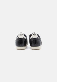Black sneakers with a reptile-embossed texture, featuring a white heel tab with logo and beige rubber outsole. Smooth design with contrasting accents.