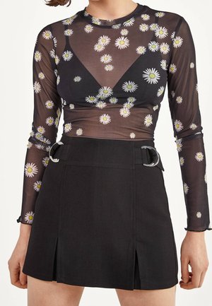 Sort mesh bluse med lange ærmer med hvidt og gult margueritprint, kombineret med et sort nederdel med høj talje med sølvspænder.