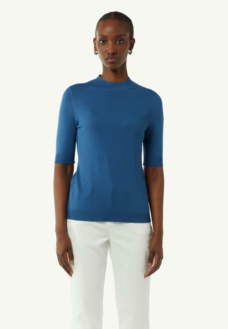 Niebieski sweter z krótkim rękawem, dziany, z okrągłym dekoltem i prążkowanymi brzegami, noszony z białymi spodniami. Gładka tekstura podkreśla równomierne szycie.