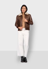 Chaqueta bomber de cuero marrón con cuello de borrego crema, cierre de cremallera al frente y dos bolsillos laterales, combinada con pantalones anchos blancos y botas negras.