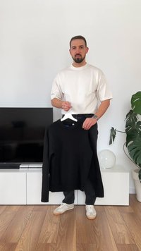 Černý pletený svetr s kulatým výstřihem, zavěšený na bílém ramínku. Má dlouhé rukávy a strukturovaný žebrovaný lem.