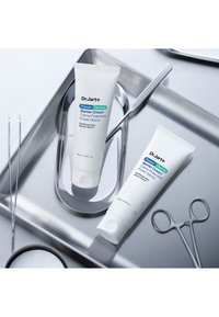 Kaksi valkoista Dr. Jart+ Power Derma Barrier Cream -voideputkea, joissa sinistä ja vihreää tekstiä, asetettuna hopeiselle tarjottimelle metallityökalujen kanssa.