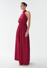 Vestido largo en tela burgundy rica, diseño de cuello halter, cintura ceñida y falda fluida. Textura suave con acentos mínimos de herrajes.