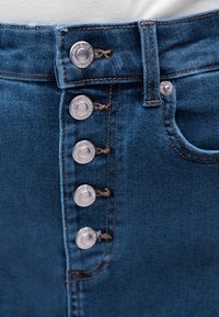 Nærbilde av blå denimjeans med fem metallknapper og oransje søm rundt linningen og lommen.