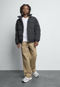 Barn med dreadlocks iført en svart boblejakke, hvit skjorte, beige bukser og hvite joggesko som står på grått gulv mot en lys vegg.