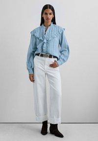 Blusa texturizada de color azul claro con un cuello con volantes y botones en la parte delantera, combinada con pantalones de pierna ancha blancos y botines de ante marrón oscuro.