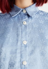 Svetle modrá blůza s květinovou texturou, límečkovým výstřihem a dekoračními bílými knoflíky ve tvaru květin s kamínkovými akcenty.