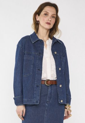 Donna che indossa una giacca e una gonna di denim blu a righe sottili sopra una camicia bianca, accessoriata con anelli d'oro, braccialetti, orecchini a cerchio e una cintura marrone.