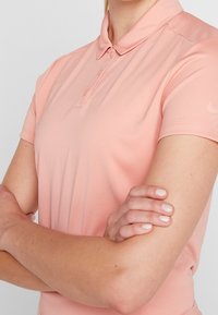 Polokošile světle růžové barvy z hladké látky, s klasickým límečkem, zapínáním na knoflíky a malým logem na rukávu.