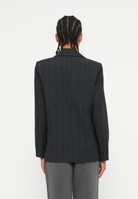Blazer à carreaux sombre avec épaules structurées, manches longues et poignets à trois boutons. Tissu texturé avec une finition lisse.