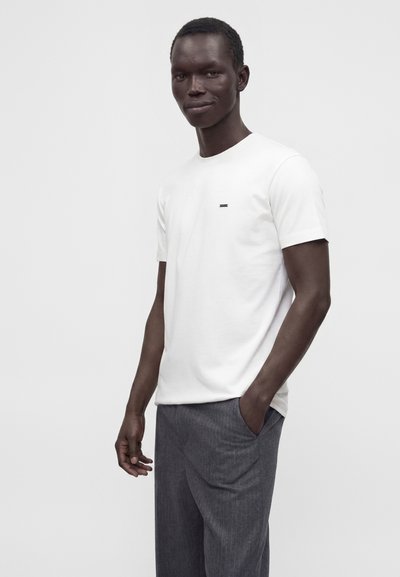Homme portant un t-shirt blanc uni à manches courtes et un pantalon gris, debout avec une main dans la poche sur un fond gris clair.