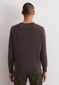 Mann mit kurzen Haaren, der einen langen braunen Pullover und dunkle Hosen trägt, steht vor einer schlichten weißen Wand.