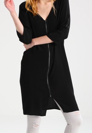 Kobieta ubrana w czarną sukienkę do kolan z przednim zamkiem błyskawicznym, rękawami 3/4 oraz białe, podarte obcisłe jeansy na jednolitym tle.