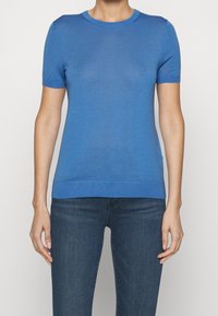 Persona con una camiseta de punto azul ajustada de manga corta y vaqueros oscuros, mostrada desde el cuello hasta las caderas sobre un fondo blanco liso.