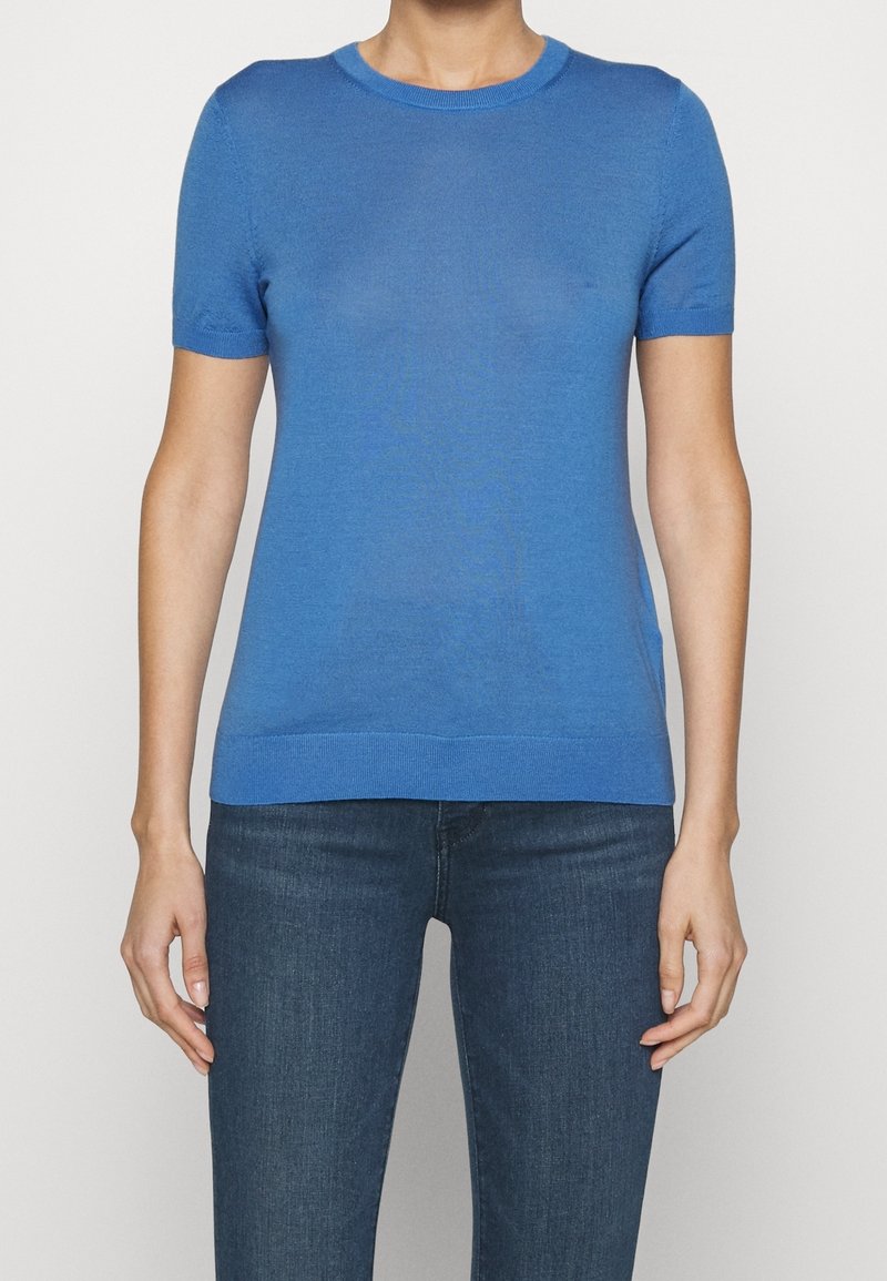 Persona con una camiseta de punto azul ajustada de manga corta y vaqueros oscuros, mostrada desde el cuello hasta las caderas sobre un fondo blanco liso.