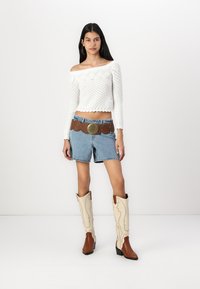 Fehér kötött, vállat szabadon hagyó crop top, kék farmer rövidnadrág, barna öv kerek csattal, bézs és barna cowboy csizma.