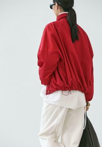 Personne aux cheveux foncés attachés en queue de cheval, portant une veste rouge ample, une chemise blanche et un pantalon blanc, portant un grand sac noir, vue de dos.