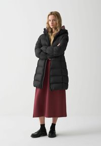 Melnā pūkainā mētelis ar dūņu rakstu, kapuci un priekšpusi ar rāvējslēdzēju. Nēsāts virs bordo kleitas, kopā ar melnām kurpēm un zeķēm.