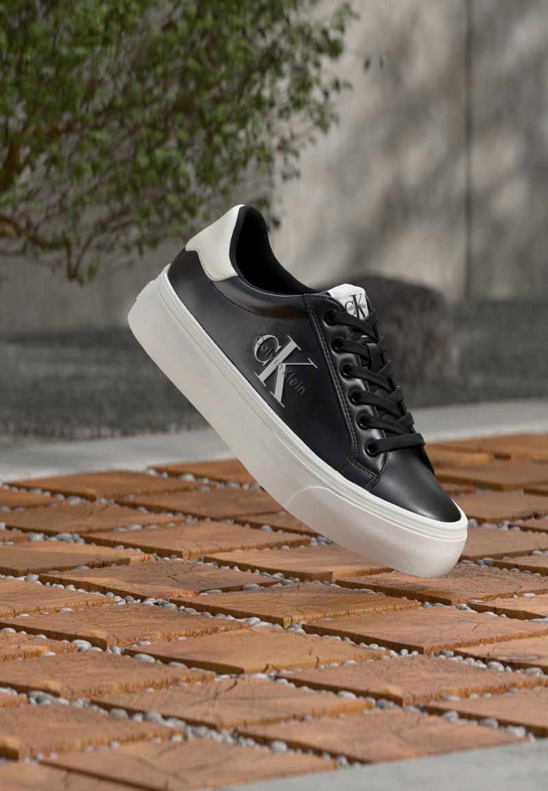 Schwarz-weißer Calvin Klein Sneaker mit Logo an der Seite, schwebend über einem Boden aus Holz- und Steinkacheln im Freien.