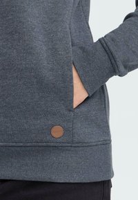 Sweatshirt gris avec une poche, des poignets côtelés et un écusson de marque brun. Fabriqué en tissu doux avec une coupe décontractée.