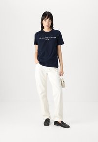 Tamsiai mėlynas medvilninis marškinėliai su balta „TOMMY HILFIGER EST. 1985“ logotipu, derinami su baltais kelnėmis ir juodais loaferiais. Paprastas dizainas, kasdienis pritaikymas.