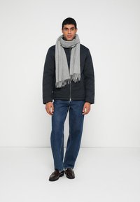 Quilted navy jacket, gray fringed scarf, blue denim jeans, and dark shoes. Model stands upright against a plain white background.