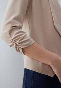 Světle béžový top vyrobený z měkké látky s nařasenými třičtvrtečními rukávy a rovný lem, nošený s tmavým denimem.