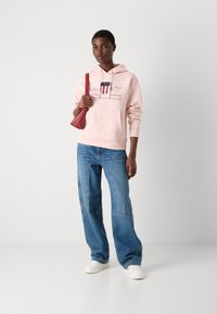 Lichtroze hoodie met een voorzak en een grafische logo, gecombineerd met blauwe wijde jeans en witte sneakers, terwijl ze een rode mini-tas vasthoudt.