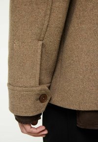 En brun ullrock har en rundad manschett med en brun knapp. Sömnadsdetaljer är synliga på tyget. Logotypen är präglad nära manschetten.