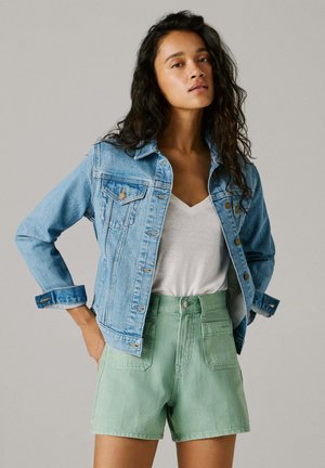 Woman with long dark curly hair wearing a light blue denim jacket, white V-neck T-shirt, and high-waisted light green shorts, standing with hands on hips.