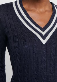 Granatowy sweter wykonany na drutach z wzorem w warkocze, z głębokim dekoltem w kształcie litery V oraz paskowaną, prążkowaną stójką w białym kolorze. Widoczna teksturowana tkanina.