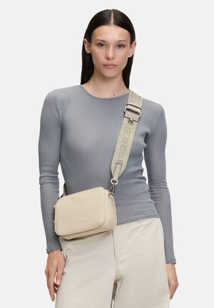 Frau mit langen dunklen Haaren trägt ein graues geripptes Langarmshirt, beige Hose und eine beige Umhängetasche mit breitem Riemen über der Schulter.