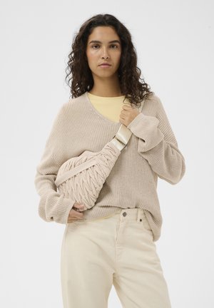Young woman with curly hair wearing beige knit sweater and pants, holding a textured beige crossbody bag against a plain white background.