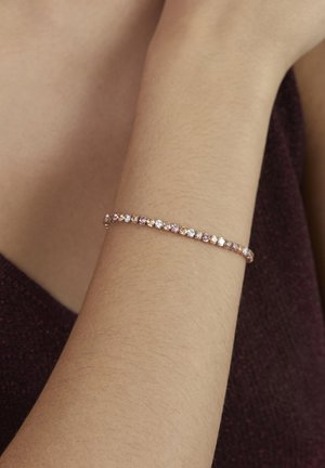 Bracciale delicato con pietre preziose alternate rosa e trasparenti indossato su un polso su uno sfondo di tessuto viola scuro.