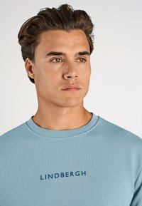 Ljusblå crewneck sweatshirt med en marinblå "LINDBERGH" logga på bröstet. Tyget verkar slätt med en enhetlig yta.