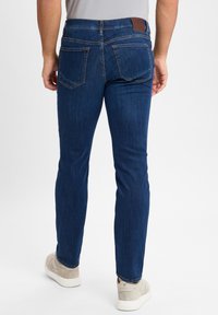 Tmavě modré slim-fit džíny s hladkou texturou. Mají koženou nášivku na pase a dvě zadní kapsy s kontrastním prošitím.